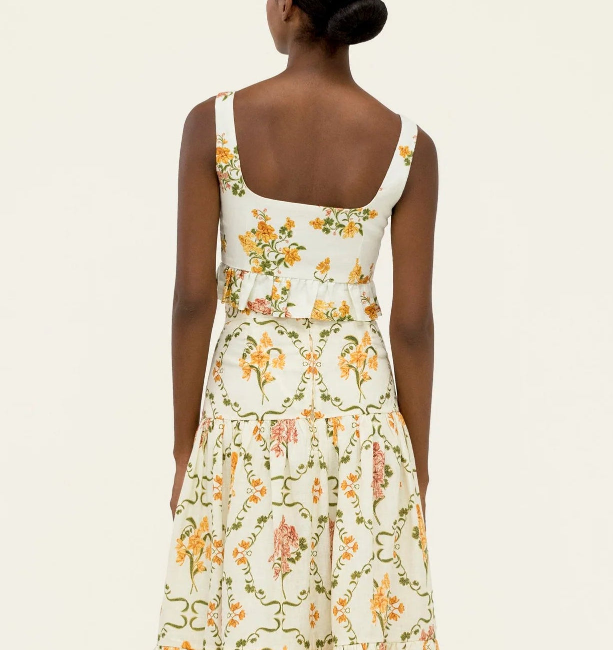 A woman faces away, wearing the Eslabon Top by Agua By Agua Bendita—a sleeveless, cream top with a square neckline and yellow, orange, and green floral pattern. Her hair is styled in a low bun.