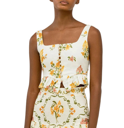 A person wearing the Eslabon Top by Agua By Agua Bendita—a sleeveless, square-neck white crop top with embroidered floral print and ruffle detail—paired with a matching floral skirt.