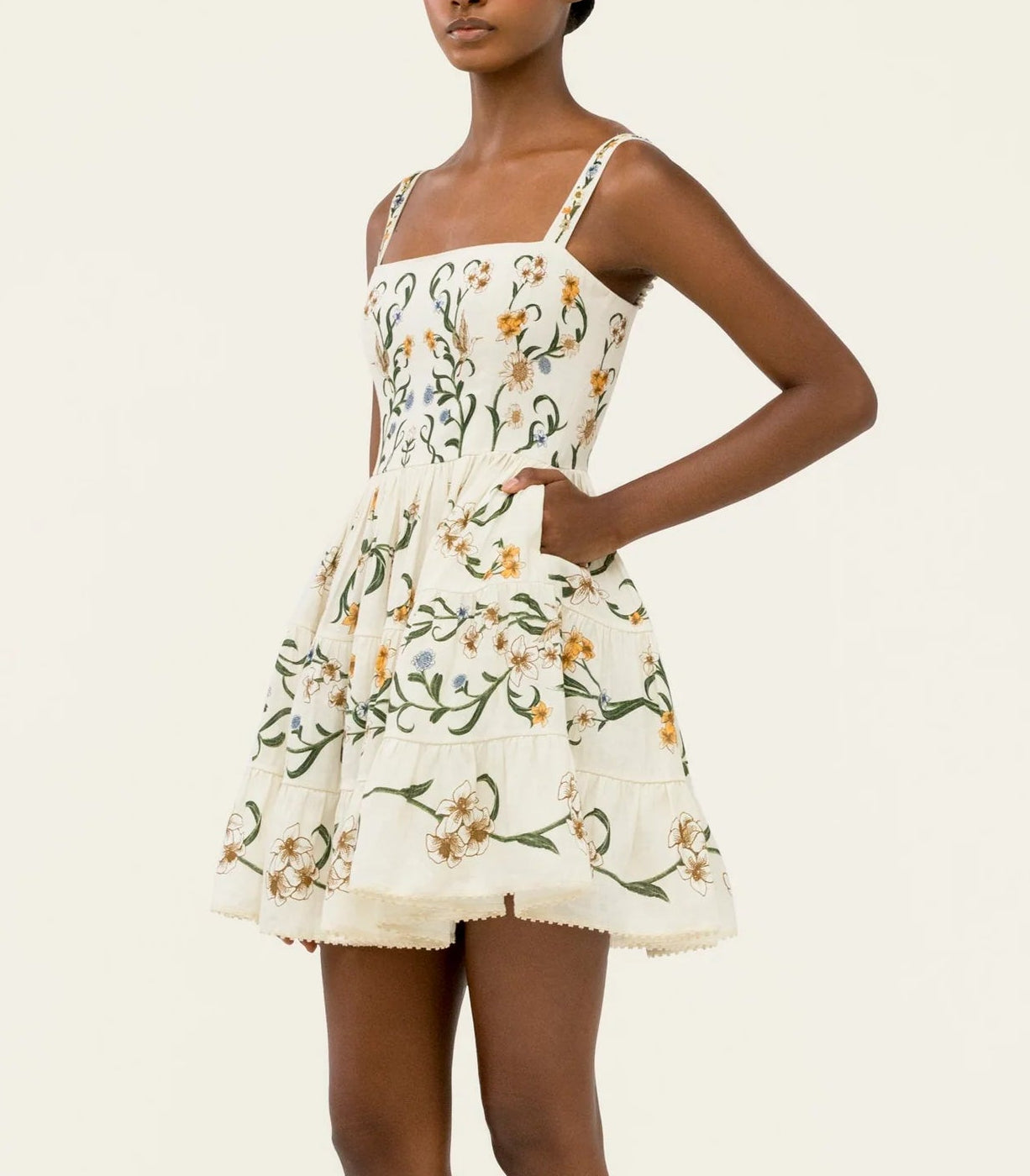 A woman wears the Lima Mini Dress by Agua By Agua Bendita, an A-line, cream cotton mini dress with floral patterns and pockets, standing against a plain light background.