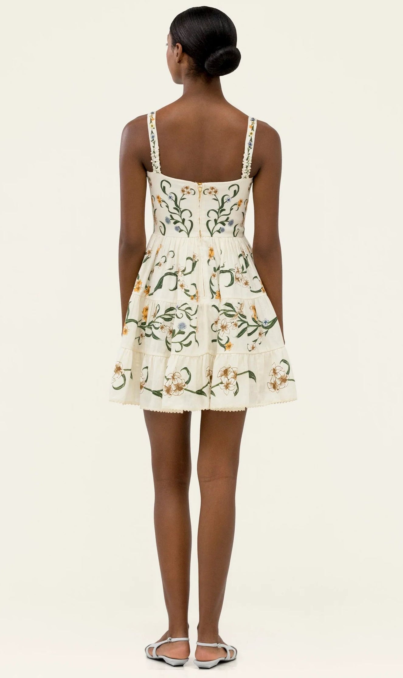 A woman faces away, wearing the Lima Mini Dress by Agua By Agua Bendita—a sleeveless, cream mini cotton dress with floral embroidery and an A-line silhouette. She pairs it with sandals and has her hair in a neat low bun.