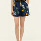 A person wears Agua By Agua Bendita’s Maratea Short, a navy two-piece set with handmade colorful floral embroidery on the top and high-waisted shorts, paired with light-colored flat shoes.