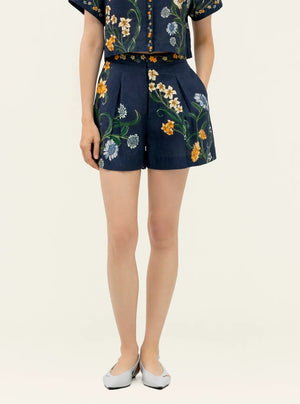 A person wears Agua By Agua Bendita’s Maratea Short, a navy two-piece set with handmade colorful floral embroidery on the top and high-waisted shorts, paired with light-colored flat shoes.