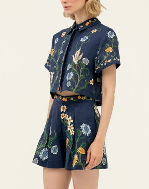 A woman wears the Virtuosa Cropped Shirt by Agua By Agua Bendita, a navy blue linen piece with handmade multicolored floral and bird embroidery, paired with matching shorts, standing against a plain background.