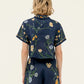 A person with shoulder-length wavy blonde hair is shown from the back, wearing an Agua By Agua Bendita Virtuosa Cropped Shirt in navy blue with a daisy print.