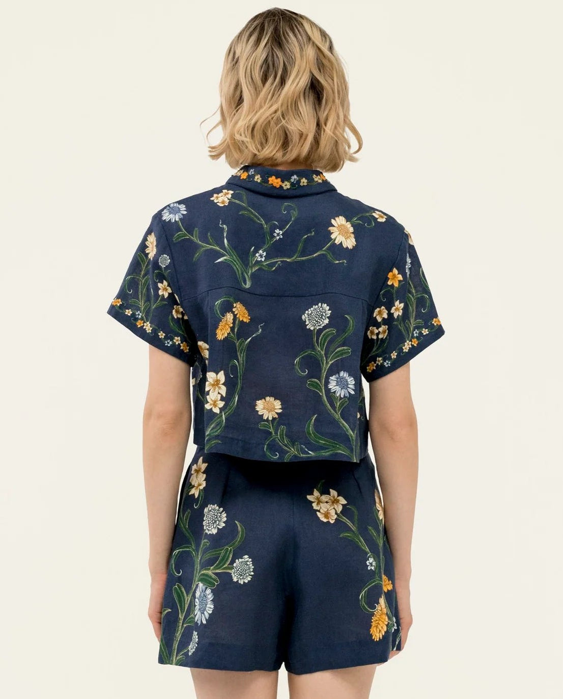 A person with shoulder-length wavy blonde hair is shown from the back, wearing an Agua By Agua Bendita Virtuosa Cropped Shirt in navy blue with a daisy print.