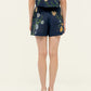 Person stands facing away from the camera, wearing Agua By Agua Bendita's Maratea Short set—a navy blue floral top and high-waisted shorts—with flat sandals against a plain light background.