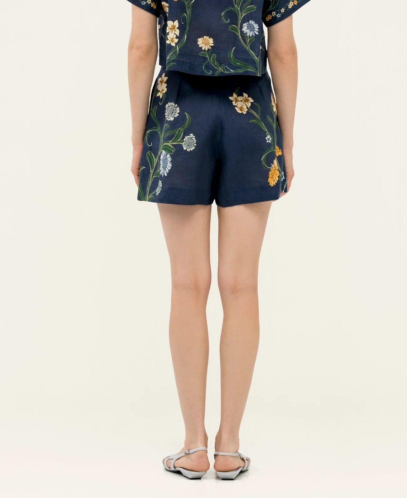 Person stands facing away from the camera, wearing Agua By Agua Bendita's Maratea Short set—a navy blue floral top and high-waisted shorts—with flat sandals against a plain light background.