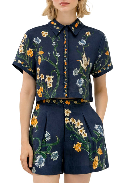 A woman wears the Virtuosa Cropped Shirt by Agua By Agua Bendita, a navy blue linen top with handmade embroidery, paired with matching high-waisted floral shorts.