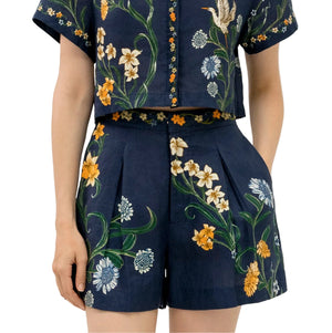 Wearing the Maratea Short by Agua By Agua Bendita, a navy blue floral matching set with pleated high-waisted shorts and a short-sleeve cropped top on illustrated fabric.