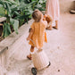 A toddler in an orange dress joyfully pushes an Olli Ella Rattan Original Luggy along a sandy path, surrounded by large green plants.