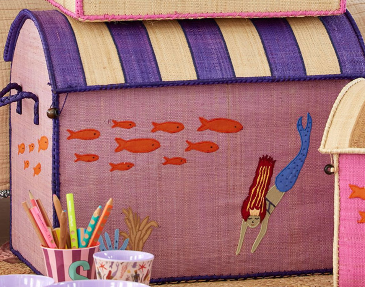 The Rice Raffia Toy Basket, adorned with purple stripes and crafted from natural materials, showcases a design of mermaids and fish. Close by, handmade storage baskets from Madagascar by the brand Rice hold cups filled with various colored pencils.