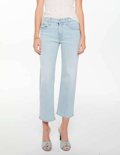 A person stands against a plain white background wearing Mother’s Mid Rise Rambler Zip Ankle Fray jeans, a sleeveless white top, and light gray open-toe heeled shoes.
