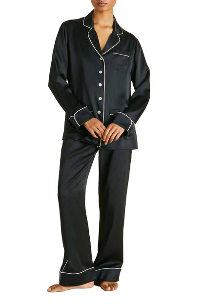 A person stands barefoot against a plain background, wearing the Olivia von Halle Coco Silk Pajamas—a luxurious black satin set with white piping, featuring a long-sleeve button-up top and matching pants evocative of an elegant evening.