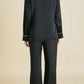 Facing away from the camera, a person models the Olivia von Halle Coco Silk Pajamas—an elegant evening set featuring a long-sleeve black satin top and matching pants, both accented with white trim—against a plain beige background.