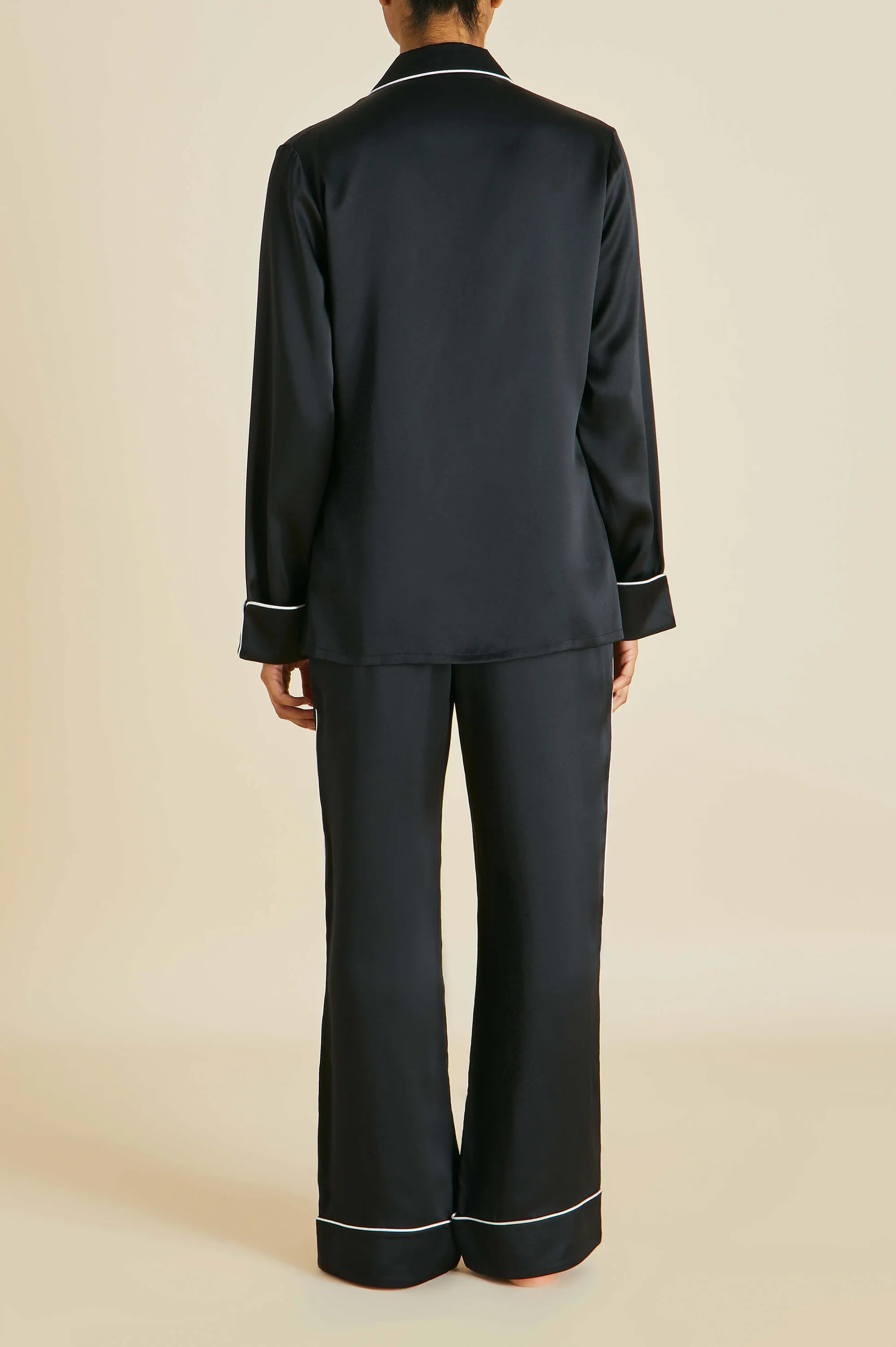 Facing away from the camera, a person models the Olivia von Halle Coco Silk Pajamas—an elegant evening set featuring a long-sleeve black satin top and matching pants, both accented with white trim—against a plain beige background.
