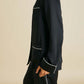A person models the Olivia von Halle Coco Silk Pajamas—a black set with white piping—against a plain beige background. The refined look captures the elegance of an evening in classic silk sleepwear, though the upper body appears partially out of frame.