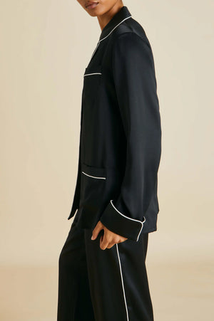 A person models the Olivia von Halle Coco Silk Pajamas—a black set with white piping—against a plain beige background. The refined look captures the elegance of an evening in classic silk sleepwear, though the upper body appears partially out of frame.