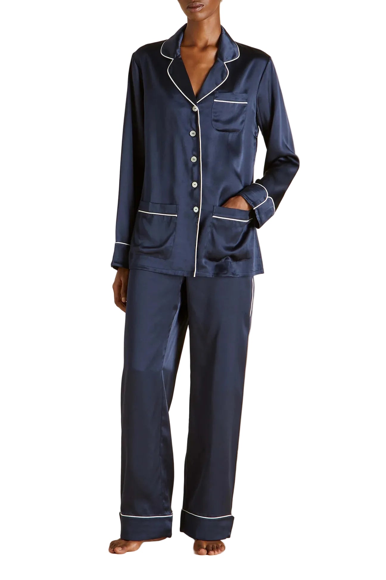 Wearing the Olivia von Halle Coco Silk Pajamas, this person models a navy silk set featuring distinctive white piping, a button-up long-sleeve top, and full-length pants. Standing barefoot against a plain background, they showcase an elegant evening look that effortlessly blends classic style with luxurious comfort.
