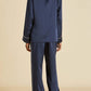 A person stands with their back to the camera, dressed in Olivia von Halle’s Coco Silk Pajamas—a matching navy set featuring a long-sleeve top and wide-leg pants accented with white piping—set against a beige background for an effortlessly elegant evening look.