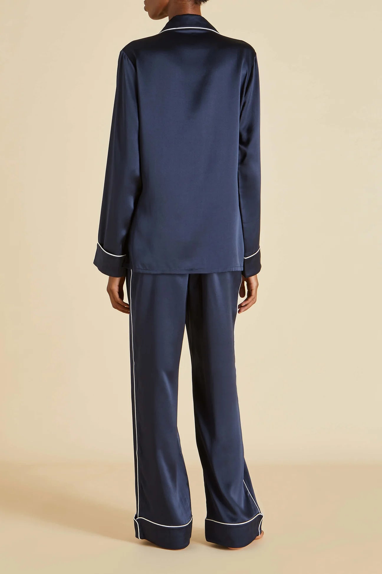 A person stands with their back to the camera, dressed in Olivia von Halle’s Coco Silk Pajamas—a matching navy set featuring a long-sleeve top and wide-leg pants accented with white piping—set against a beige background for an effortlessly elegant evening look.