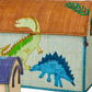 The Rice Raffia Toy Basket features a green dinosaur on the lid and a blue stegosaurus on the side, adding playful charm. A smaller matching basket is partially visible, perfect for fun and practical storage.