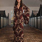 A woman stands in a stable aisle wearing the Johanna Ortiz One Palm Only Tunic Dress—a floor-length style with flared sleeves and sandals, showcasing an intricate brown and beige design inspired by the Equine Mola motif. Sunlight streams through an open door behind her.