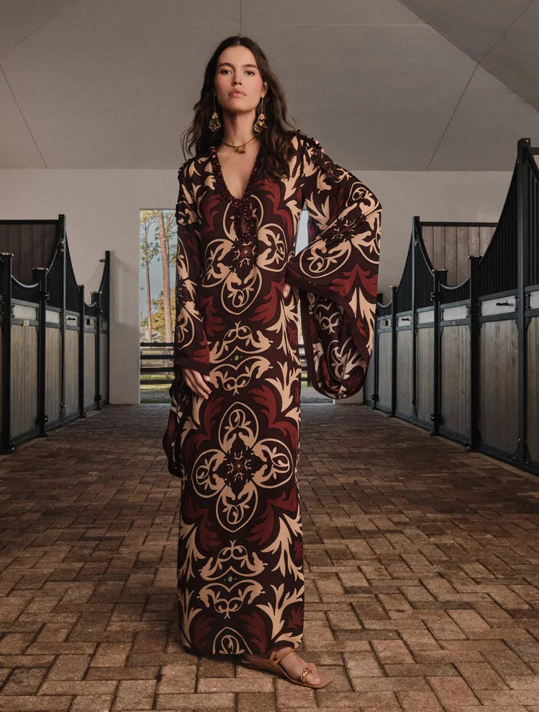 A woman stands in a stable aisle wearing the Johanna Ortiz One Palm Only Tunic Dress—a floor-length style with flared sleeves and sandals, showcasing an intricate brown and beige design inspired by the Equine Mola motif. Sunlight streams through an open door behind her.