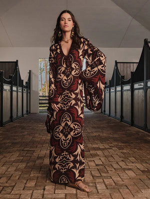 A woman stands in a stable aisle wearing the Johanna Ortiz One Palm Only Tunic Dress—a floor-length style with flared sleeves and sandals, showcasing an intricate brown and beige design inspired by the Equine Mola motif. Sunlight streams through an open door behind her.