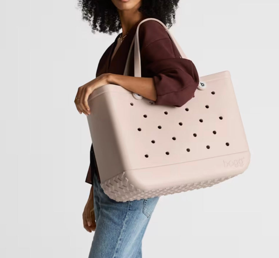 A woman carries an Original Bogg® Bag tote by Bogg Bags in light pink, featuring signature holes and woven details at the bottom, while wearing a brown top and blue jeans.