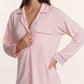 A woman stands against a plain background wearing the Eberjey Gisele Long PJ Set—a light pink, long sleeve button-front top with chest pocket and collar, paired with matching straight leg pants.