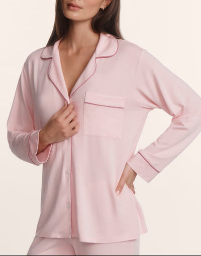 A woman stands against a plain background wearing the Eberjey Gisele Long PJ Set—a light pink, long sleeve button-front top with chest pocket and collar, paired with matching straight leg pants.