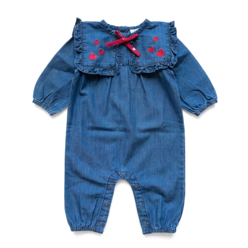 The Baby Girls’ Celiciane Romper by Louis Louise features blue chambray fabric with long sleeves, a sailor collar adorned with a ruffled edge and red bow, and charming small red heart prints on the chest; it also offers a convenient snap inseam for easy changing.