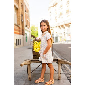 A young girl wearing the Louis Louise Girls’ Lola Terry Heart Overall stands by a wooden cart filled with yellow apples on a city sidewalk.