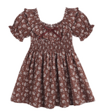 The Girls’ Dillion Dress by Rylee & Cru features a brown fabric with a white floral print, short puff sleeves, a ruffle neckline, smocked bodice, and is accented with a small maroon bow at the neckline.