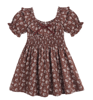 The Girls’ Dillion Dress by Rylee & Cru features a brown fabric with a white floral print, short puff sleeves, a ruffle neckline, smocked bodice, and is accented with a small maroon bow at the neckline.