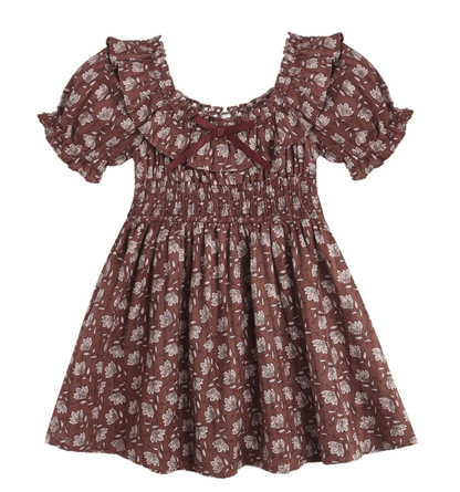 The Girls’ Dillion Dress by Rylee & Cru features a brown fabric with a white floral print, short puff sleeves, a ruffle neckline, smocked bodice, and is accented with a small maroon bow at the neckline.