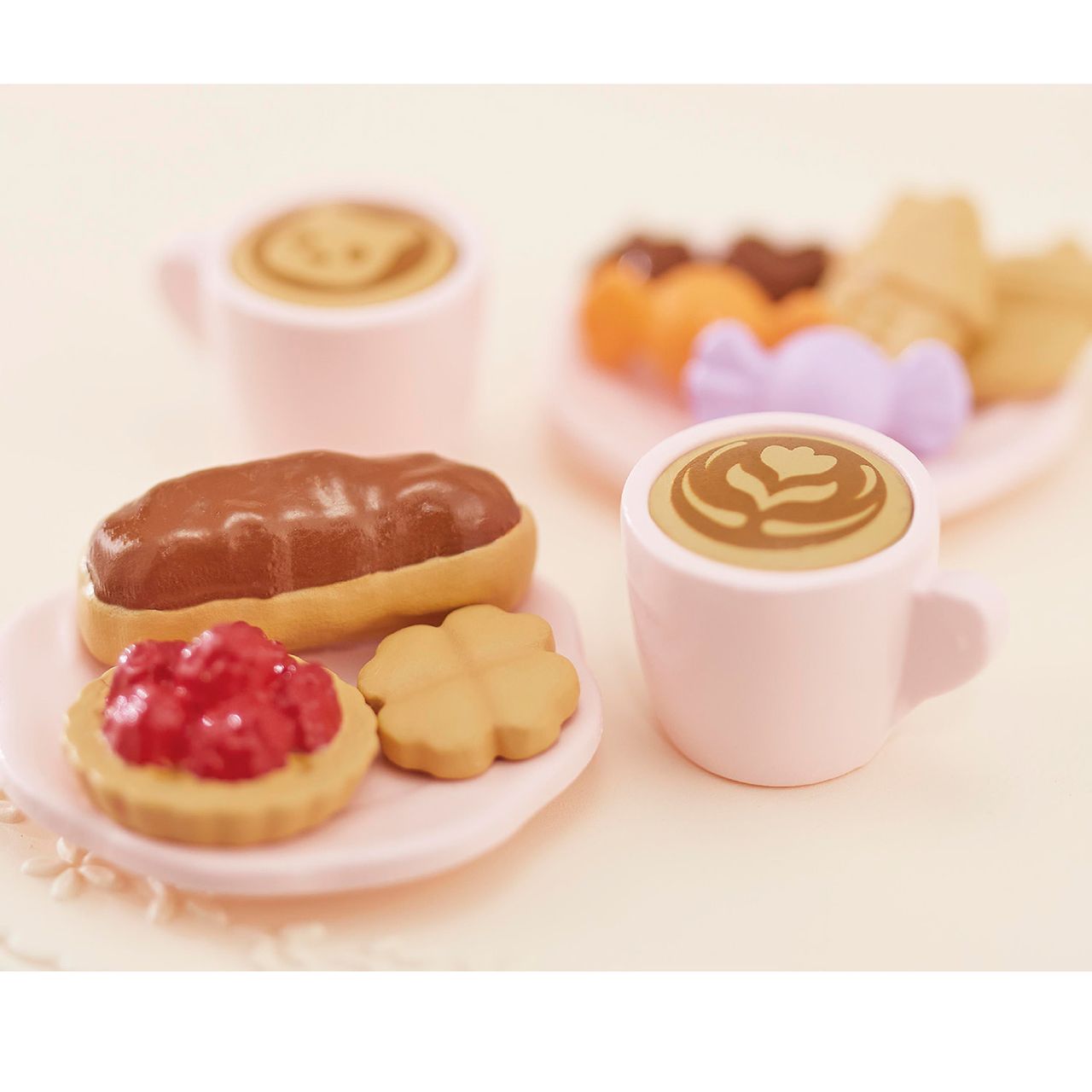 The Calico Critters Sweets Party Set features two pink coffee cups with latte art and two plates filled with an éclair, tart, cookies, and assorted sweets, all arranged on a light surface—perfect for imaginative playtime or pairing with your favorite teatime accessories.