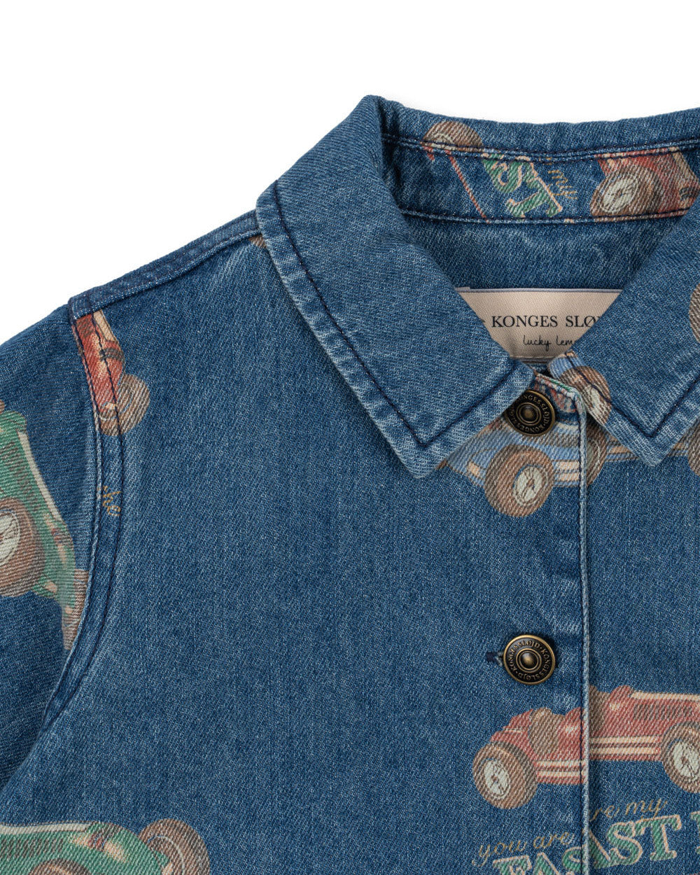 Close-up of the Konges Slojd Baby Boys' and Boys' Magot Jacket—a blue denim shirt with collar and buttons, featuring a vintage car print and brand label for a touch of playful charm to any look.