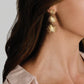 A person wearing Anabel Aram Palm Triple Fan Dangle Earrings, featuring 18K gold-plated stacked, star-shaped triple fan palm charms.