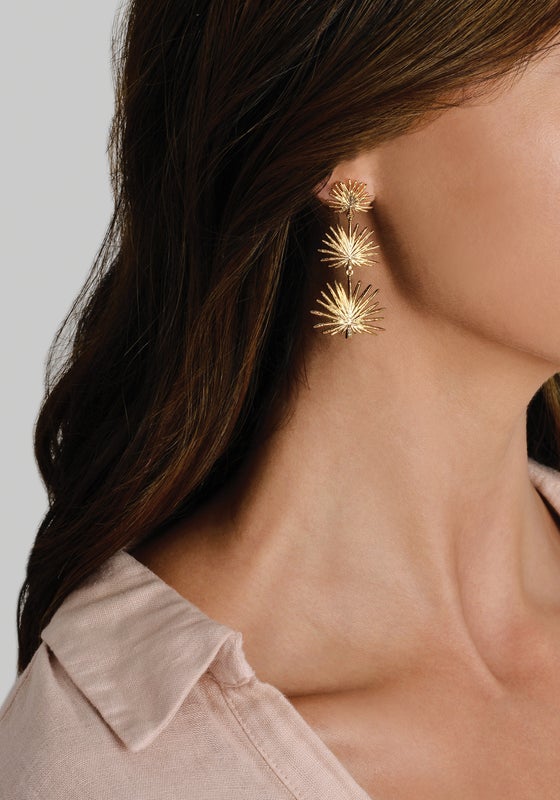 A person wearing Anabel Aram Palm Triple Fan Dangle Earrings, featuring 18K gold-plated stacked, star-shaped triple fan palm charms.