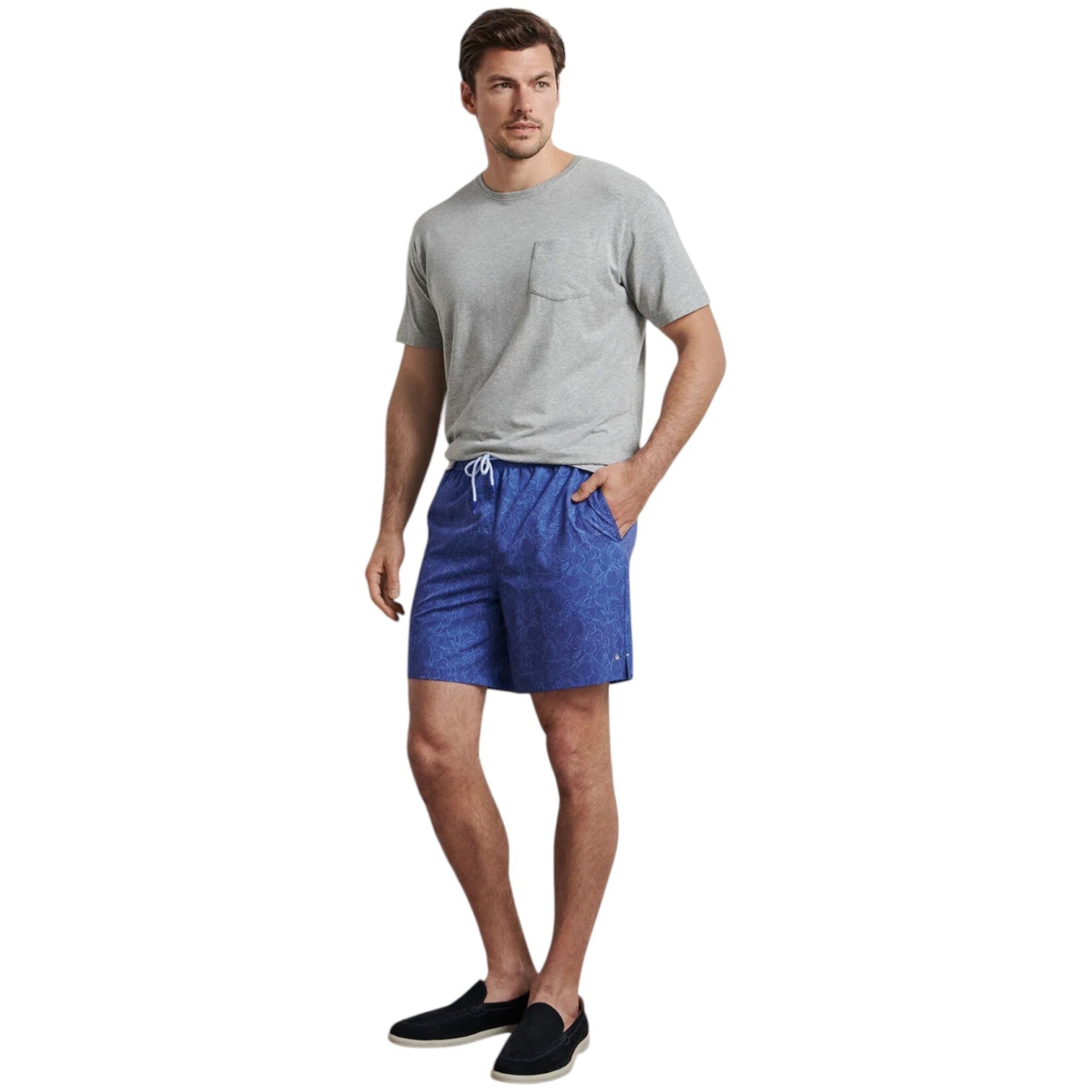 A man wears a grey t-shirt, Peter Millar Paloma Swim Trunk with four-way stretch and a subtle pattern, and black slip-on shoes, with one hand in his pocket.