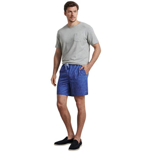 A man wears a grey t-shirt, Peter Millar Paloma Swim Trunk with four-way stretch and a subtle pattern, and black slip-on shoes, with one hand in his pocket.