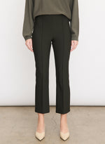 A person stands against a plain background, wearing vince’s Niki Mid Rise Crop Flare Pant in black Italian cotton-blend with pintuck legs, paired with a long-sleeve olive top and beige pointed-toe shoes. Only the lower body is visible.