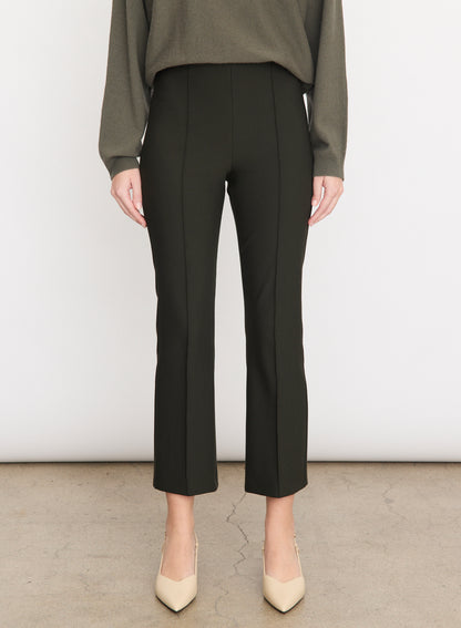 A person stands against a plain background, wearing vince’s Niki Mid Rise Crop Flare Pant in black Italian cotton-blend with pintuck legs, paired with a long-sleeve olive top and beige pointed-toe shoes. Only the lower body is visible.