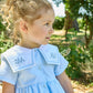 A young girl with curly blonde hair wears the Kidiwi Baby Girls' and Girls' Paola Dress, featuring a sky blue piqué fabric and white embroidered collar, as she sits outdoors surrounded by trees and greenery.