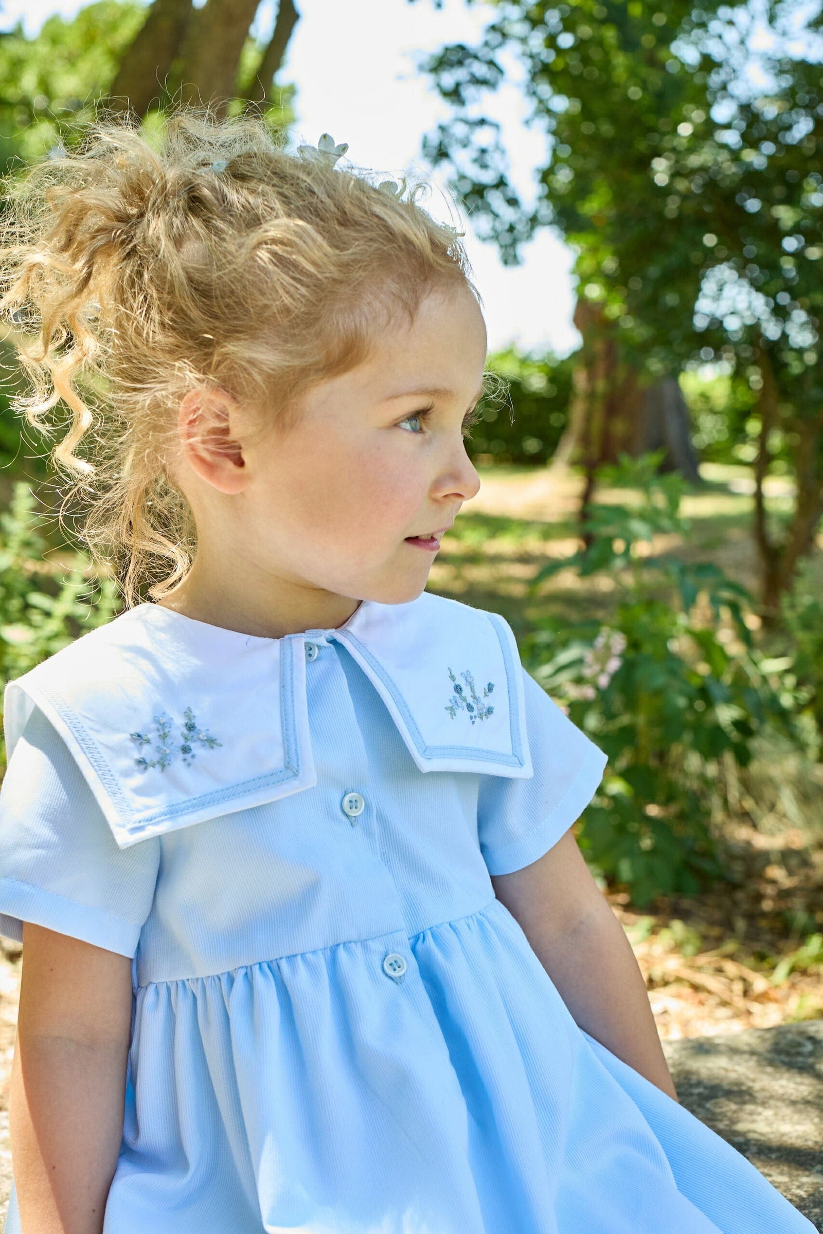 A young girl with curly blonde hair wears the Kidiwi Baby Girls' and Girls' Paola Dress, featuring a sky blue piqué fabric and white embroidered collar, as she sits outdoors surrounded by trees and greenery.