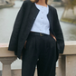 A person stands on a stone bridge wearing the POSSE Rio Top—a white buttoned blouse—paired with a relaxed fit black suit jacket and high-waisted black trousers. A river and trees are visible in the background.