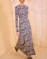 A person in the Ulla Johnson Naia Midi Dress with long sleeves and ikat print stands indoors against a beige textured background with hanging fringe decor.