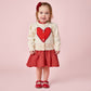 A young child stands against a pink background wearing the Pink Chicken Baby Girls' Constance Sweater, featuring a large red heart, paired with a red skirt, matching shoes, and a bright red bow in her hair.