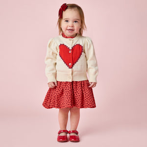 A young child stands against a pink background wearing the Pink Chicken Baby Girls' Constance Sweater, featuring a large red heart, paired with a red skirt, matching shoes, and a bright red bow in her hair.